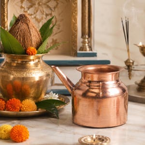 Puede incluir: Un recipiente de cobre pulido con un pico lateral se encuentra junto a una olla de latón que contiene un coco y hojas verdes. La olla de latón está adornada con flores de caléndula naranjas y amarillas. Se ven una vela y varitas de incienso.