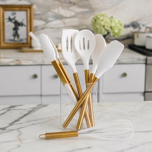 May include: A set of white silicone kitchen utensils with gold-toned metal handles, including tongs, a slotted turner, a slotted spoon, a solid spoon, and a spatula, all stored in a clear glass container, with a matching whisk lying in front.