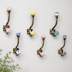 May include: Six wall-mounted coat hooks made of antique brass metal, each featuring two ceramic knobs. The knobs display various patterns and colors, including red floral, yellow polka dot, blue floral, pink solid, teal geometric, and black stripes.