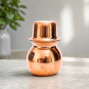 Peut inclure: Un récipient en cuivre poli avec un corps arrondi et un col étroit, surmonté d'un couvercle assorti en forme de tasse inversée. La surface métallique présente une teinte brun-orangé chaude avec des stries horizontales et un fini brillant.