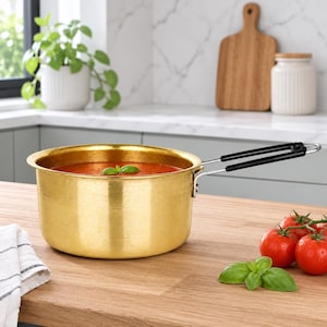 May include: A round, metallic gold-coloured saucepan with a long, black-handled metal extension. The pot is filled with a smooth, vibrant red tomato-based sauce, garnished with a single green basil leaf floating on the surface of the liquid.