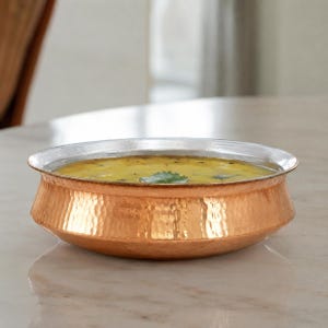 May include: A round, shallow serving bowl with a hammered copper exterior and a smooth silver-toned metal rim. The bowl is filled with a yellow, textured liquid containing small dark specks and a single green herb leaf floating on the surface.