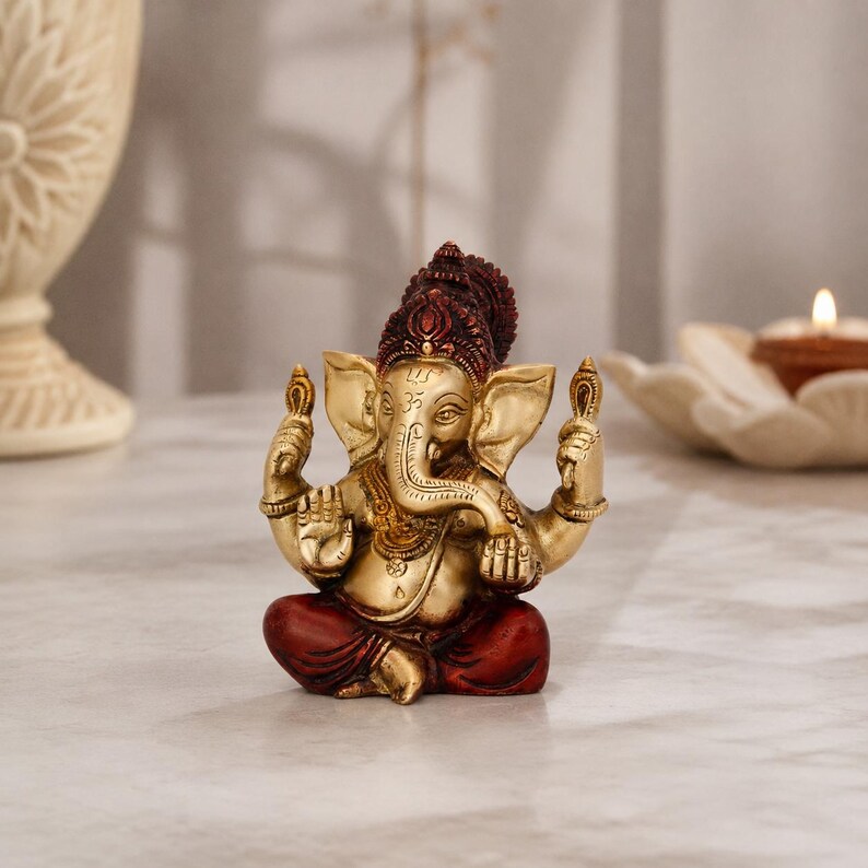 May include: A small Ganesha figurine featuring a metallic gold finish on the head, torso, and arms, with deep red coloring on the headdress and lower body. The deity is depicted in a seated position with four arms, holding objects in the upper hands.