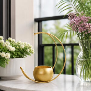 Peut inclure: Un bougeoir en métal doré composé d'une base en forme de bol sphérique soutenue par une structure fine en forme de croissant. L'objet présente une finition métallique lisse, avec un arc incurvé s'élevant de la base vers une petite sphère.