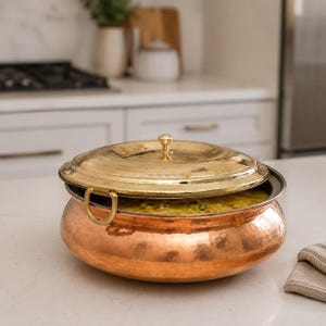 May include: A round, hammered copper serving bowl with a wide base and two side handles. The bowl is filled with a yellow, textured food dish and is partially covered by a polished, hammered brass lid featuring a small, central knob handle.