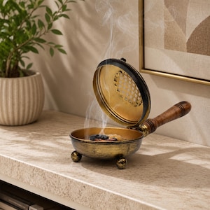 May include: A round, metallic incense burner with a distressed gold finish, three small spherical feet, and a hinged lid featuring a grid pattern. A wooden handle extends from the side, and thin wisps of white smoke rise from the interior.