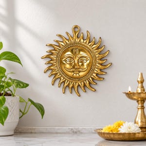 Puede incluir: Un adorno de pared circular de metal dorado que presenta un rostro de sol estilizado con bigote y una marca en la frente. El sol tiene numerosos rayos ondulados que irradian hacia afuera y una pequeña anilla en la parte superior.