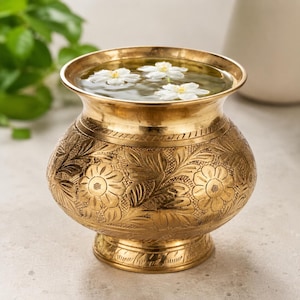Puede incluir: Un cuenco de latón pulido con un borde ancho y acampanado y un cuerpo redondeado con intrincados motivos florales y de hojas grabados a mano. El recipiente está lleno de agua clara y contiene tres flores blancas flotando en la superficie.