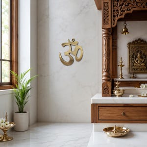 Könnte beinhalten: Ein metallisches, goldfarbenes Om-Symbol als Wandbehang mit gebürsteter Oberfläche. Das Design zeigt das traditionelle Sanskrit-Zeichen mit einem geschwungenen Oberteil, einer mittleren Schlaufe und einem unteren Schweif auf einer weißen Marmorwand.