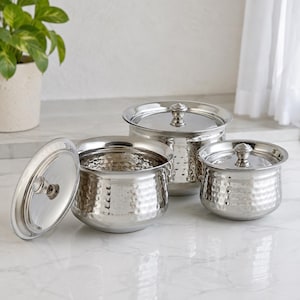 May include: A set of three silver-colored metal serving bowls with hammered textures and matching lids. Each bowl features a rounded body and a polished finish. One lid is placed upright next to the bowls, while the other two lids are on top.