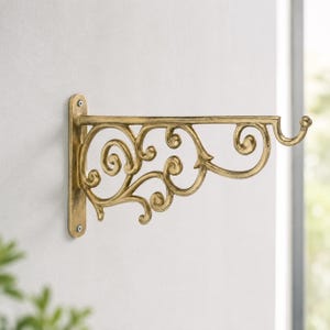 Puede incluir: Un soporte de pared de metal dorado con un diseño ornamental de volutas y un gancho curvado en el extremo. El soporte está fijado a una superficie plana con dos tornillos visibles, mostrando un acabado desgastado y patrones metálicos complejos.