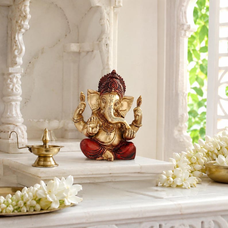 May include: A seated Ganesha statue featuring a metallic gold finish on the body and head, accented with deep red clothing and a dark red crown. The deity has four arms, with the upper hands holding small objects and the lower hands in mudra gestures.