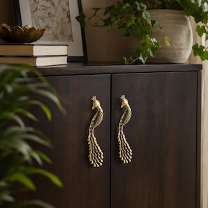 May include: Two brass-colored cabinet handles shaped like peacocks with detailed plumage and crests. The handles are mounted vertically on a dark brown wooden cabinet door, featuring a metallic finish with textured wing and tail feather patterns.