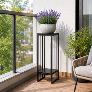 Op de afbeelding: Een hoge, zwarte metalen plantenstandaard met een vierkant blad en een onderste plank, voorzien van een verticaal stangontwerp. Een lichtgrijze, geribbelde, ronde keramische pot staat op de standaard met bloeiende, paarse lavendelplanten.