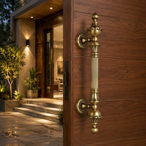 May include: A vertical brass door handle with a fluted central column and decorative finials at the top and bottom, mounted on a dark brown wood-grain door. The handle features circular mounting plates with intricate engraved detailing.