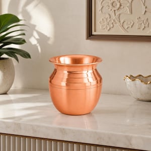 May include: A polished copper pot with a rounded body and a wide, flared rim. The vessel features a series of horizontal engraved lines encircling the upper section, creating a subtle textured detail against the smooth, metallic surface.