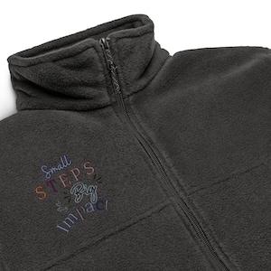 May include: A charcoal gray fleece jacket with a high collar and a full-length front zipper. The chest features embroidered text that reads Small STEPS Big Impact in a mix of purple, orange, and pink thread colors with small heart icons.