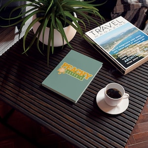 Puede incluir: Un diario de tapa dura en color verde salvia con el texto "PEACHY MIND" en una fuente estilizada de color amarillo y naranja en la portada. El diario está colocado sobre una superficie oscura de listones junto a una taza de café blanca y una revista.