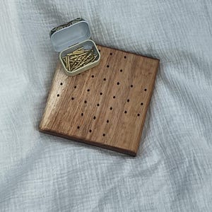 May include: A square wooden board featuring a grid of small drilled holes for peg placement. Resting on the top left corner of the board is a small, open, rectangular metal tin filled with a collection of thin, gold-coloured metal pegs.