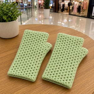 May include: A pair of light green fingerless gloves crafted with a textured crochet stitch pattern. The gloves feature a rectangular shape with a dedicated opening for the thumb, showcasing a consistent grid-like weave throughout the fabric.