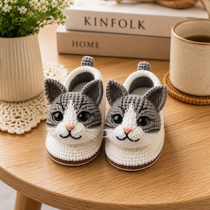Puede incluir: Un par de zapatillas de ganchillo hechas a mano con diseño de caras de gato. Las zapatillas cuentan con una base blanca, orejas y frente grises, grandes ojos negros de plástico, una pequeña nariz rosa y bigotes blancos bordados a los lados.
