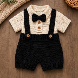 May include: A cream-colored crocheted short-sleeve shirt with a black bow tie and two brown buttons on the placket. The shirt is attached to black crocheted shorts with integrated suspender straps, each featuring a single brown button at the waist.