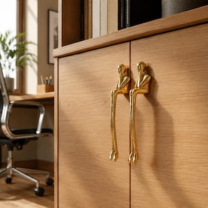 Pode incluir: Dois puxadores de armário em metal dourado polido com formato de figuras estilizadas sentadas com pernas longas estendidas para baixo. As figuras estão montadas numa porta de armário de madeira clara, com uma superfície lisa, reflexiva e forma humana.