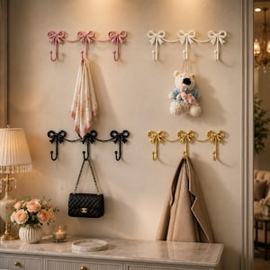 Puede incluir: Cuatro percheros de pared en rosa, blanco, negro y dorado, cada uno con tres ganchos y detalles decorativos de lazos. Los percheros están dispuestos en cuadrícula, con un paño estampado, un oso de peluche, un bolso acolchado negro y un abrigo.