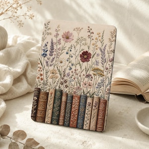 May include: A rectangular e-reader cover featuring a cream-colored background with an embroidered-style print of various wildflowers and a row of ten vertical, antique-style book spines along the bottom edge in shades of brown, green, and blue.