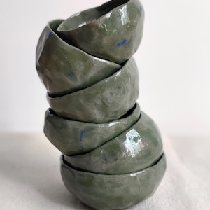 May include: A vertical stack of five handmade ceramic bowls with an irregular, organic shape. The bowls feature a glossy, sage green glaze with subtle, scattered flecks of deep blue pigment throughout the smooth, reflective surface.