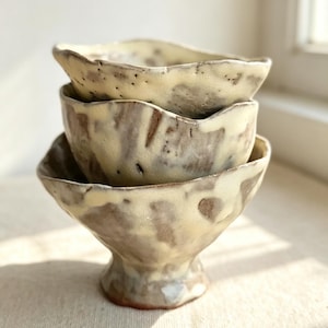 May include: A stack of two handmade ceramic bowls with an irregular, wavy rim. The bowls feature a mottled glaze with creamy off-white tones layered over a dark brown, earthy base, revealing a textured, speckled surface and a pedestal base.