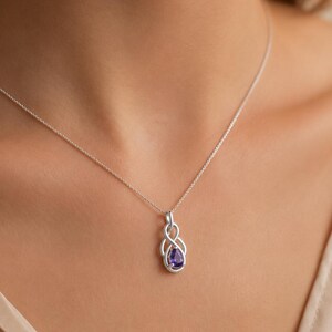 Könnte beinhalten: Eine silberne Halskette mit einem zarten Anhänger in verschlungenem Unendlichkeits-Design und einem einzelnen tropfenförmigen violetten Edelstein in der Mitte. Der Anhänger hängt an einer dünnen, polierten Silberkette am Schlüsselbein.