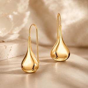 Puede incluir: Un par de pendientes colgantes de tono dorado con un colgante en forma de gota suave y pulida, suspendido de un gancho largo y curvado. La superficie metálica es muy reflectante y muestra un acabado amarillo cálido y brillante.