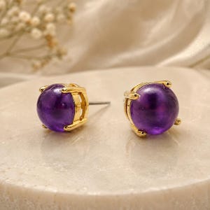 Peut inclure: Une paire de boucles d'oreilles à clous avec des pierres cabochons violettes polies et rondes, serties dans des griffes en métal doré. Chaque boucle présente une tige métallique visible à l'arrière, conçue pour les oreilles percées.