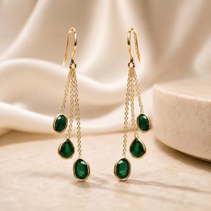 Può includere: Un paio di orecchini pendenti in oro caratterizzati da tre catenelle delicate di diversa lunghezza, ognuna terminante con una piccola pietra preziosa ovale di colore verde intenso. Gli orecchini sono appesi a semplici ganci dorati.