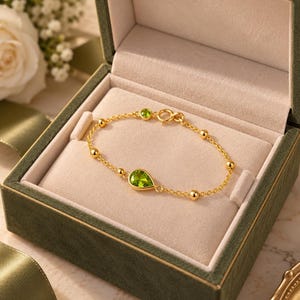 Peut inclure: Un bracelet en chaîne dorée orné d'une pierre précieuse verte en forme de poire, d'une petite pierre verte ronde et de plusieurs perles dorées lisses. Le bracelet est présenté dans un écrin à bijoux doublé de velours vert foncé.