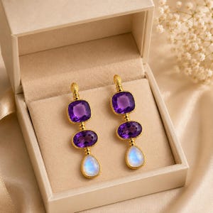 Peut inclure: Une paire de boucles d'oreilles pendantes dorées composées d'un alignement vertical de trois pierres précieuses. Chaque boucle présente une améthyste violette carrée, une améthyste ovale et une pierre de lune irisée en forme de poire.