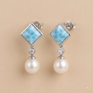 May include: A pair of silver drop earrings featuring a square-cut light blue stone, a small round clear accent stone, and a white spherical pearl dangling at the bottom. The earrings have a post-back closure and a polished metallic finish.