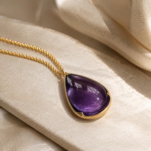 May include: A gold necklace featuring a large, teardrop-shaped purple amethyst gemstone pendant set in a smooth gold bezel. The pendant is attached to a delicate gold chain, showcasing a polished finish and a deep, vibrant violet hue.