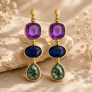 Peut inclure: Une paire de boucles d'oreilles pendantes dorées composées de trois pierres précieuses facettées superposées. La pierre supérieure est une améthyste violette carrée, celle du milieu un saphir bleu ovale et celle du bas une agate mousse verte en forme de goutte.