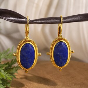 May include: A pair of gold-toned leverback earrings featuring oval-shaped, deep blue lapis lazuli stones. Each stone is encased in a textured, beaded gold frame with small decorative spheres on the sides, suspended from a curved gold hook mechanism.