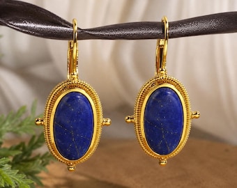 Puede incluir: Un par de pendientes dorados con cierre de palanca y piedras de lapislázuli azul intenso de forma ovalada. Cada piedra está engastada en un marco dorado texturizado con cuentas y pequeñas esferas decorativas en los laterales.