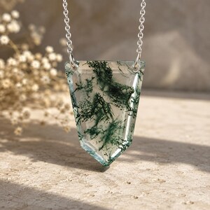 May include: A shield-shaped moss agate pendant featuring translucent stone with dark green, fern-like inclusions, suspended from a delicate silver-toned metal chain. The pendant has faceted edges and two small circular holes for the chain attachment.
