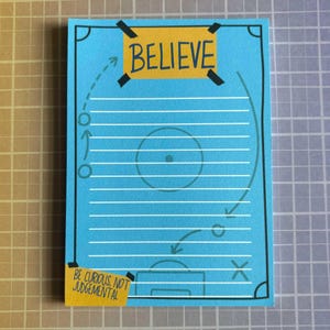 Peut inclure: Un bloc-notes rectangulaire sur fond bleu clair avec une bannière jaune en haut portant le mot BELIEVE. Le centre affiche un schéma de jeu sportif bleu foncé avec des lignes et des cercles. Une étiquette jaune indique BE CURIOUS, NOT JUDGEMENTAL.