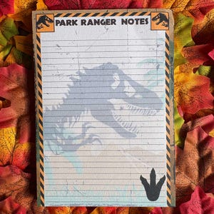 May include: A rectangular notepad titled PARK RANGER NOTES at the top, featuring a large grey dinosaur silhouette, horizontal lines, and black and yellow hazard stripes along the sides. A black dinosaur footprint icon is at the bottom right.