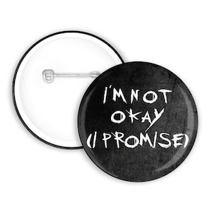 Può includere: Una spilla rotonda con uno sfondo nero usurato e il testo bianco I'M NOT OKAY (I PROMISE) scritto con un carattere irregolare in stile manuale. Il retro della spilla mostra un meccanismo a spillo in metallo argentato per l'aggancio.