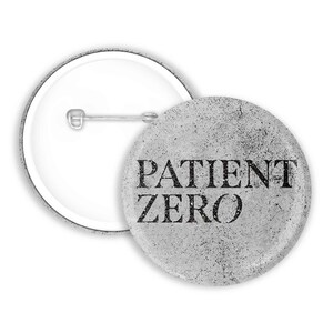 Peut inclure: Un badge rond avec un fond texturé gris clair à l'aspect vieilli, portant l'inscription PATIENT ZERO en lettres capitales noires et grasses. Le dos du badge présente un mécanisme d'épingle en métal argenté pour la fixation.