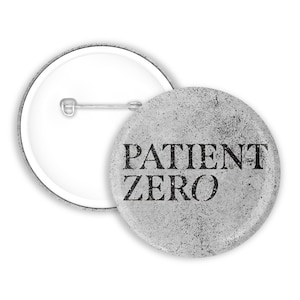 Może przedstawiać: Okrągła przypinka z postarzanym, jasnoszarym, teksturowanym tłem oraz napisem PATIENT ZERO wykonanym pogrubionymi, czarnymi wielkimi literami. Tył przypinki jest biały i posiada srebrny metalowy mechanizm z agrafką.