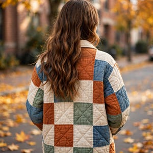 May include: A long-sleeved quilted jacket featuring a patchwork pattern of square blocks in muted shades of rust orange, sage green, and cream. The garment has a collared neckline and a visible stitched grid texture throughout the fabric.