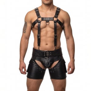May include: A black leather chest harness featuring silver-toned metal buckles, rivets, and a central O-ring, paired with matching black leather chaps that include a wide waist belt with a buckle and side pockets, branded with the Leather Empire logo.